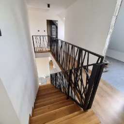 Drewniane schody z czarną, geometryczną balustradą w nowoczesnym wnętrzu, widok z góry, jasne ściany, częściowo wykończone poddasze w tle.