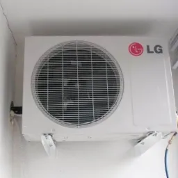 Biała jednostka zewnętrzna klimatyzatora LG zamontowana na ścianie wewnątrz pomieszczenia, widoczne przewody i uchwyty montażowe, w tle okno z żaluzjami.