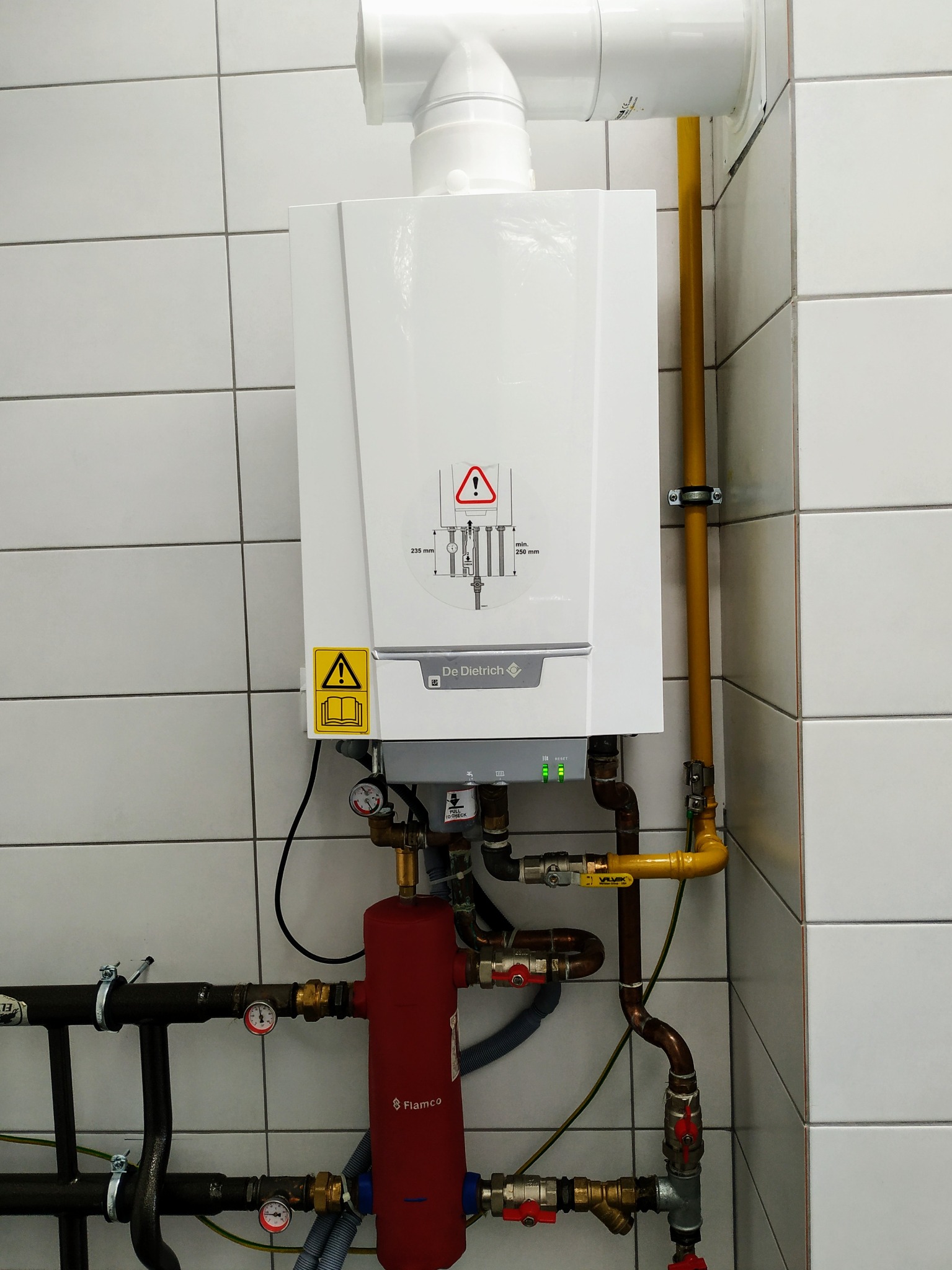 Biały, wiszący piec gazowy De Dietrich na tle białych płytek, z widocznym pionowym żółtym przewodem gazowym, czerwonym naczyniem przeponowym Flamco i licznymi rurami miedzianymi oraz zaworami.