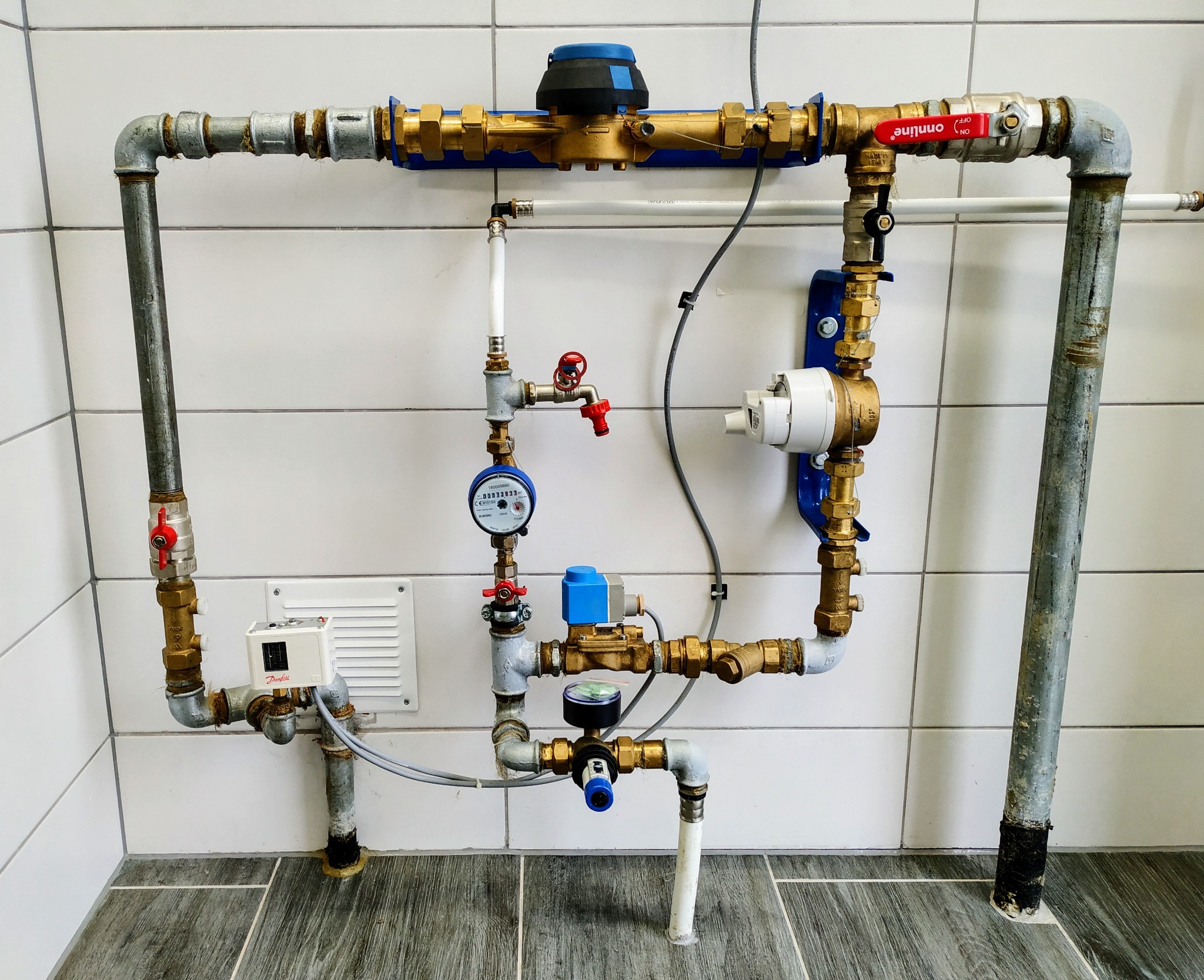 Kompleksowy system rur i zaworów z licznikiem wody, manometrem i elektronicznym regulatorem Danfoss zamontowany na ścianie z białych płytek.