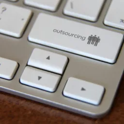 Fragment klawiatury z napisem 'outsourcing' i ikoną trzech sylwetek, leżącej na drewnianym biurku.