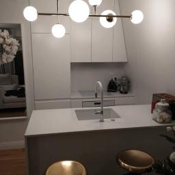 Solmar - Minimalistyczna, biała kuchnia z wyspą, oświetlona nowoczesną lampą z kulistymi kloszami, dwa złote hokery przed wyspą, w tle lustro z kwiatową dekoracją.