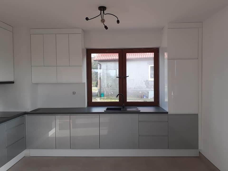 Nowoczesna kuchnia z szarymi, lakierowanymi frontami szafek dolnych i białymi górnymi, czarnym blatem i oknem z brązową ramą, widok na budynek z zewnątrz.