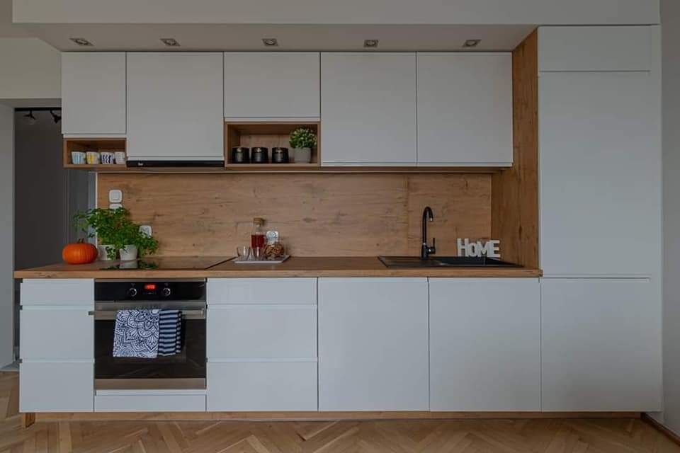 Nowoczesna kuchnia z białymi szafkami, drewnianym blatem i backsplash'em, z dynią i napojem na blacie, oraz tekstylną ściereczką na piekarniku.