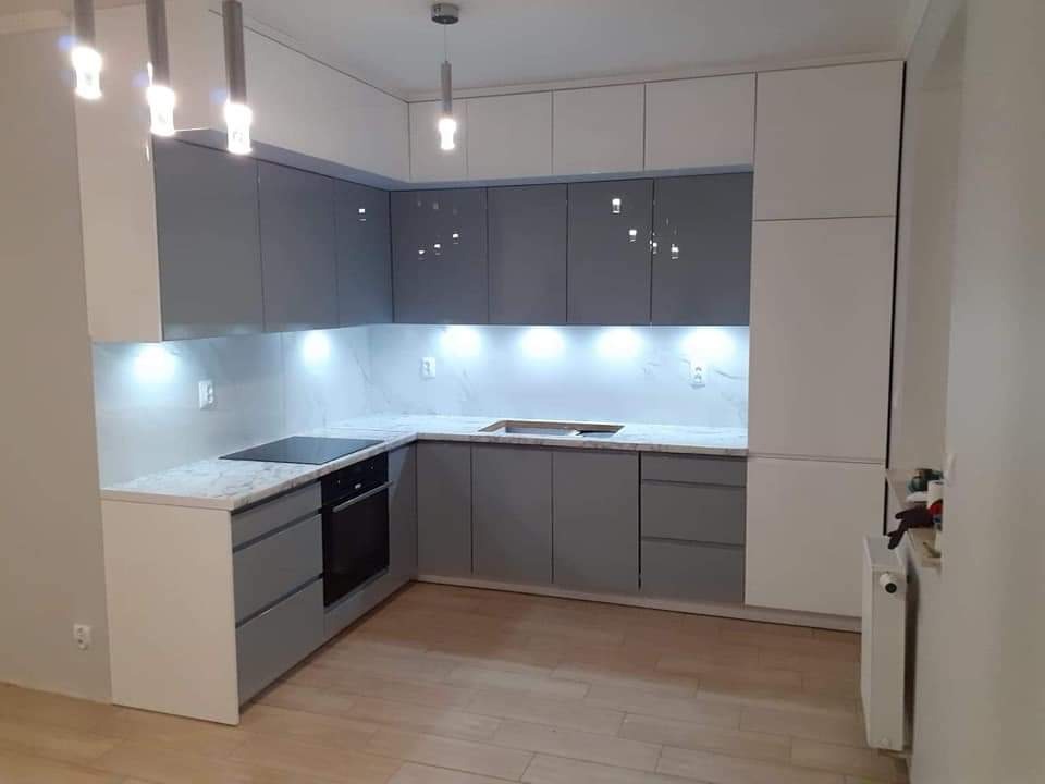 Nowoczesna, narożna kuchnia w odcieniach szarości i bieli, z podświetlanymi szafkami górnymi i marmurowym blatem, oświetlona wiszącymi lampami, z widoczną płytą indukcyjną i piekarnikiem.