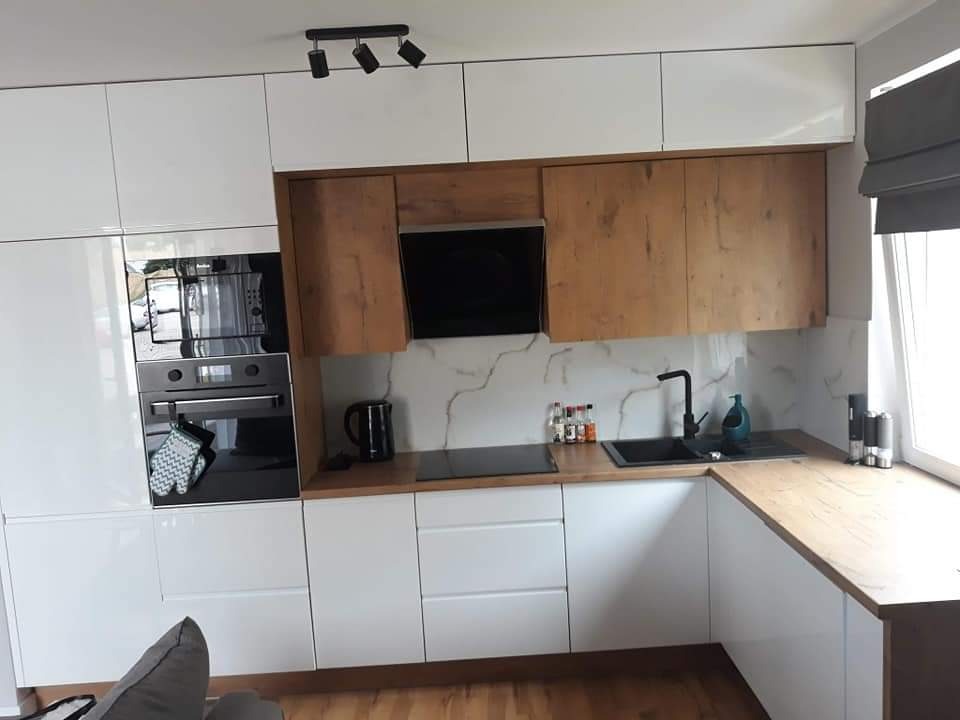 Nowoczesna kuchnia z białymi, błyszczącymi frontami dolnych szafek i drewnopodobnymi górnymi szafkami, czarną armaturą i marmurowym backsplash'em, oświetlona trzema reflektorami punktowymi.