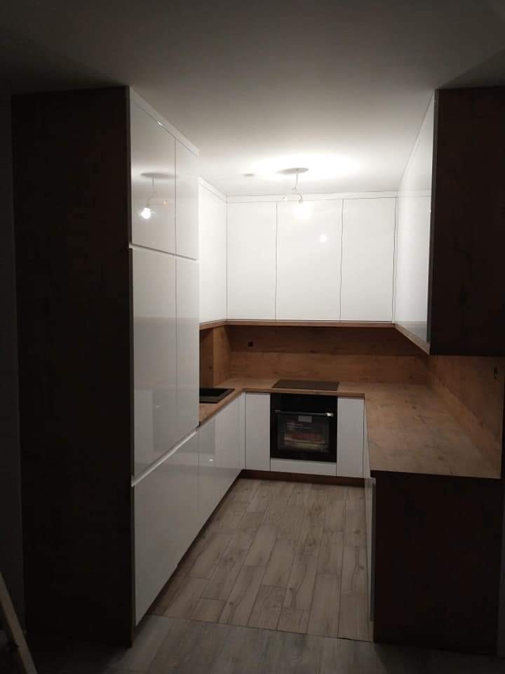 Nowoczesna kuchnia w kształcie litery U z białymi, lakierowanymi frontami i drewnianym blatem. Ciemnobrązowe elementy wykończeniowe tworzą kontrast z jasnymi powierzchniami. Widoczna płyta...