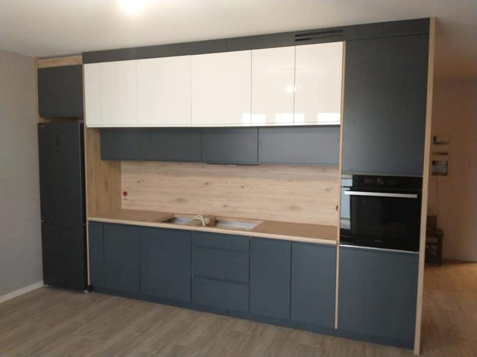 Nowoczesna zabudowa kuchenna z szarymi szafkami dolnymi i białymi górnymi, drewnianym blatem oraz panelem ściennym w odcieniu jasnego drewna, z wbudowanym piekarnikiem i lodówką.