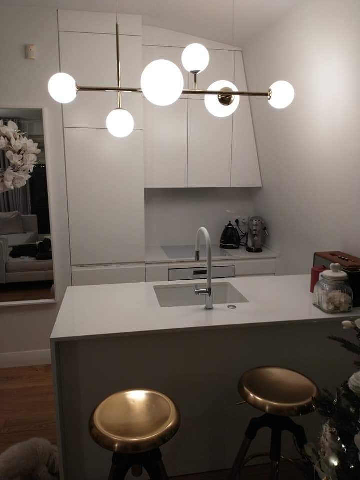 Minimalistyczna, biała kuchnia z wyspą, oświetlona nowoczesną lampą z kulistymi kloszami, dwa złote hokery przed wyspą, w tle lustro z kwiatową dekoracją.