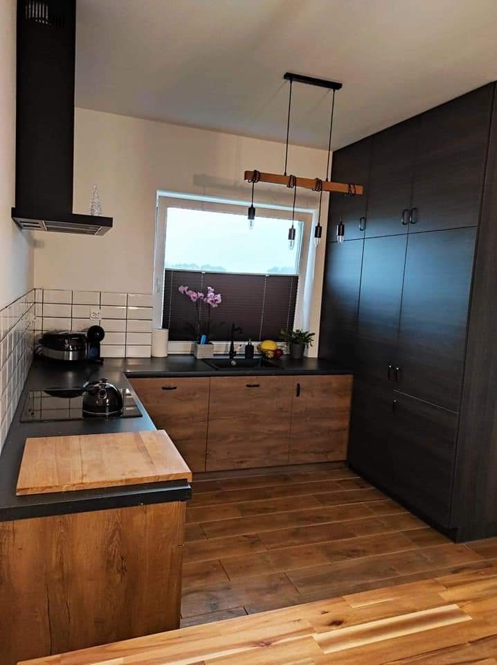 Nowoczesna kuchnia z drewnianymi frontami szafek dolnych, czarnym blatem i wysoką zabudową w odcieniach grafitu, oświetlona wiszącą lampą z drewnianym elementem, z drewnianą podłogą i białymi...