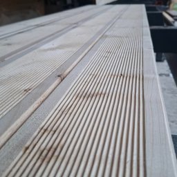 SERVISCO - Deska z wyfrezowanymi rowkami, jasne drewno, ułożona poziomo, widoczne detale obróbki, w tle maszyna stolarska, perspektywa z dołu.