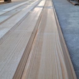 SERVISCO - Bliski kadr na ułożone deski z jasnego drewna z widocznym rysunkiem słojów, w tle maszyny stolarskie i worki z trocinami w warsztacie.