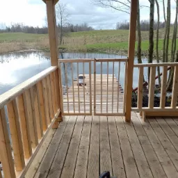 Drewniana altana z widokiem na jezioro i pomost, świeżo po budowie, z widocznym kompresorem na podłodze.