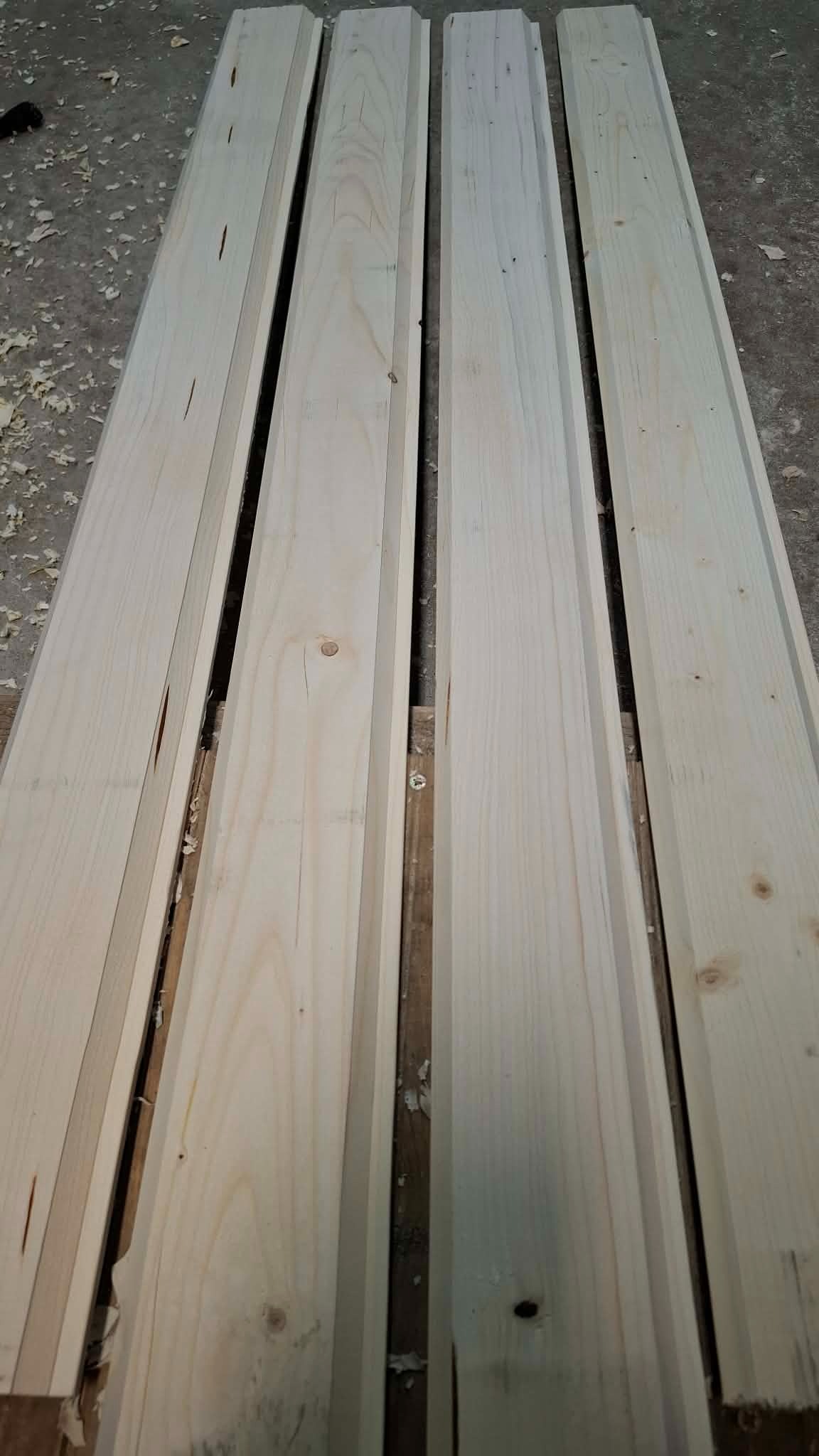 Cztery surowe, jasne deski z widocznymi słojami drewna, ułożone równolegle na drewnianej palecie, z drobnymi wiórami w tle. Deski przygotowane do dalszej obróbki.