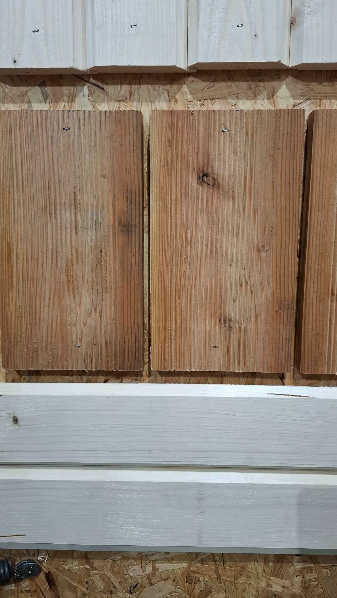 Próbki drewna: trzy surowe deski z widocznymi słojami i dwie pomalowane na biało, prezentowane na płycie OSB. Widoczne gwoździe mocujące.