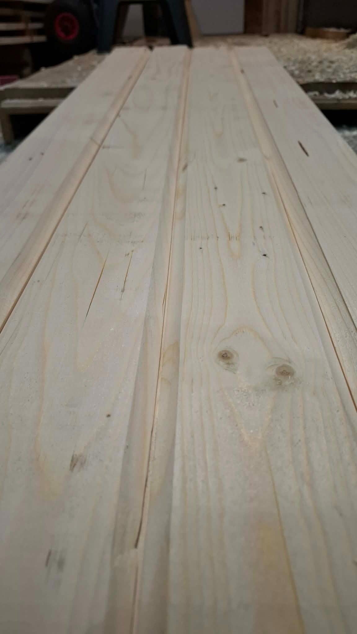 Jasne, surowe deski drewniane ułożone równolegle, widoczne sęki i delikatne usłojenie, perspektywa zbieżna, w tle fragment wózka i resztki obróbki drewna.