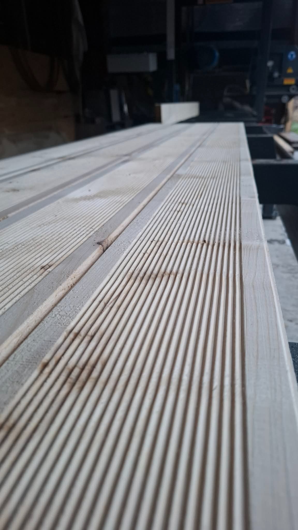 Deska z wyfrezowanymi rowkami, jasne drewno, ułożona poziomo, widoczne detale obróbki, w tle maszyna stolarska, perspektywa z dołu.