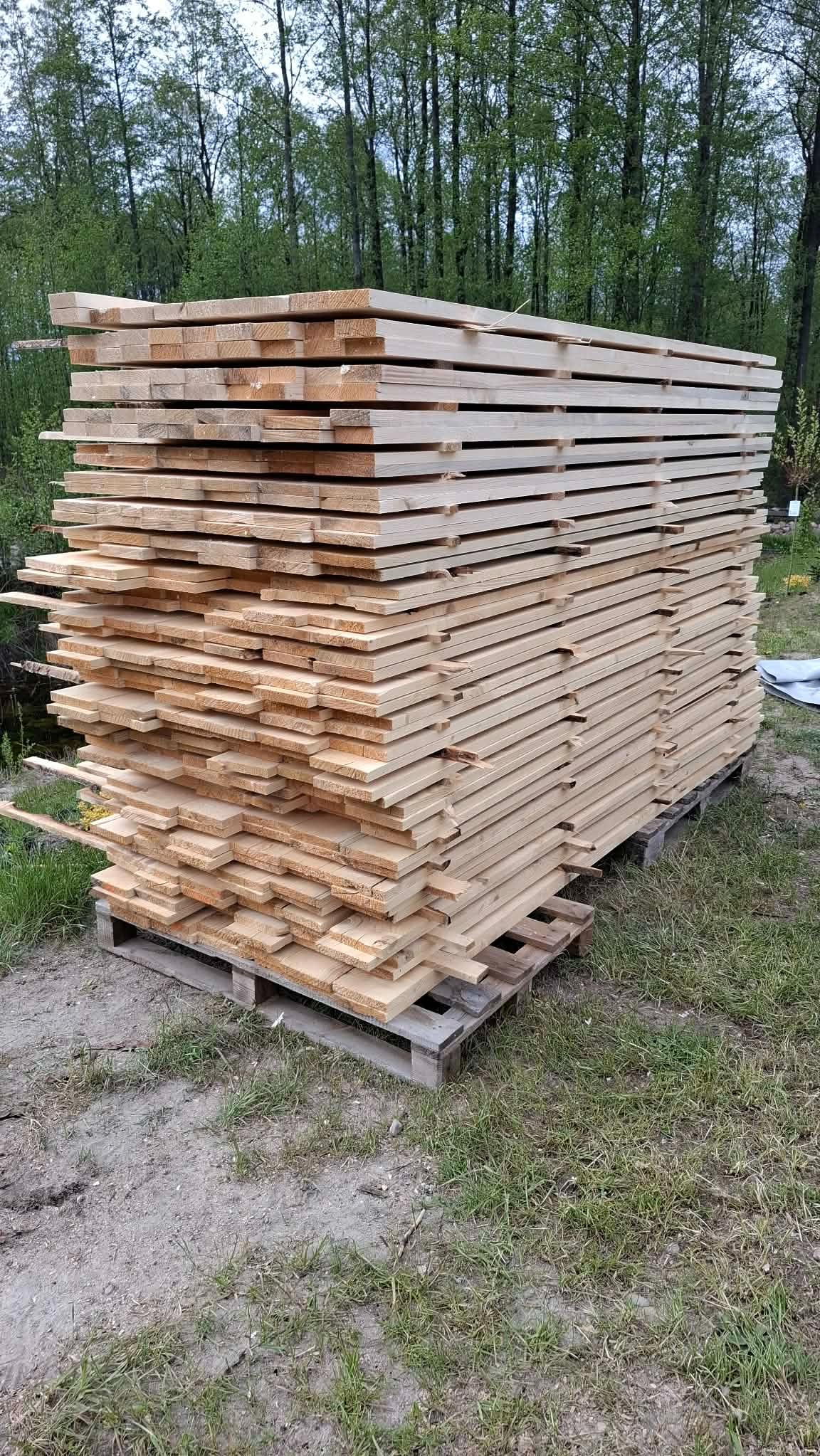 Stos desek ułożonych na palecie, widoczny na tle zieleni drzew. Deski jasne, świeżo przetarte, ułożone równo. Paleta stoi na ziemi porośniętej trawą.