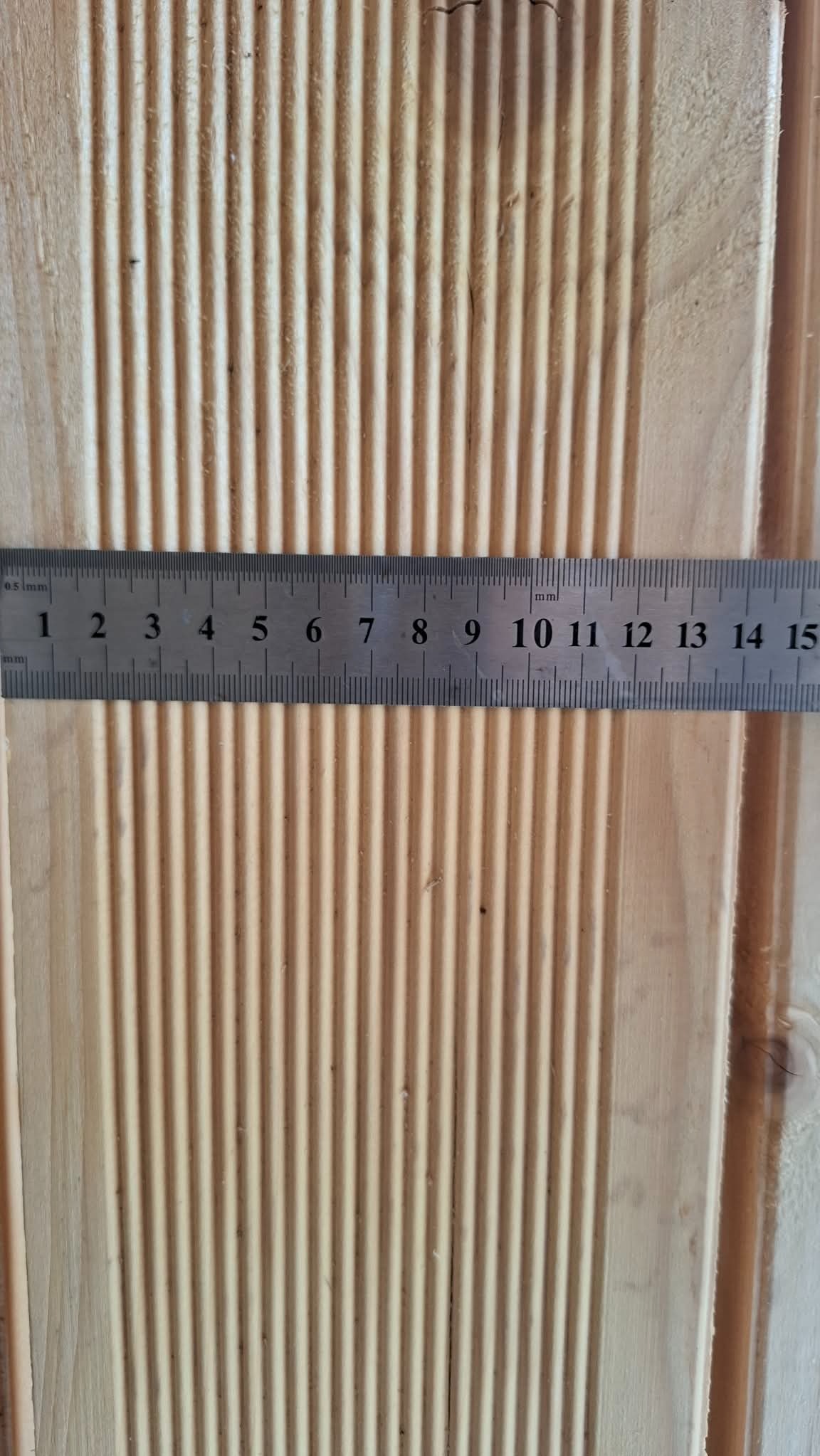 Deska boazeryjna z widocznym profilem, mierzona metalową linijką. Jasne drewno, pionowe żłobienia. Pomiar szerokości deski.