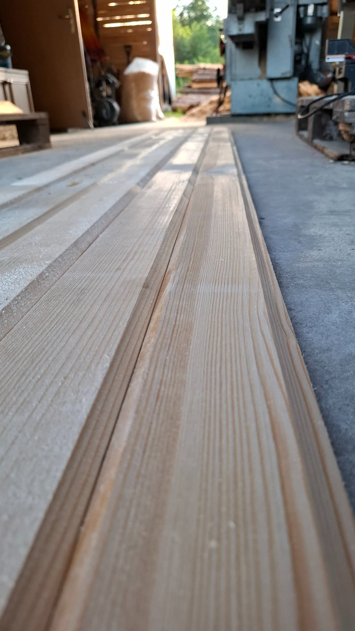 Bliski kadr na ułożone deski z jasnego drewna z widocznym rysunkiem słojów, w tle maszyny stolarskie i worki z trocinami w warsztacie.