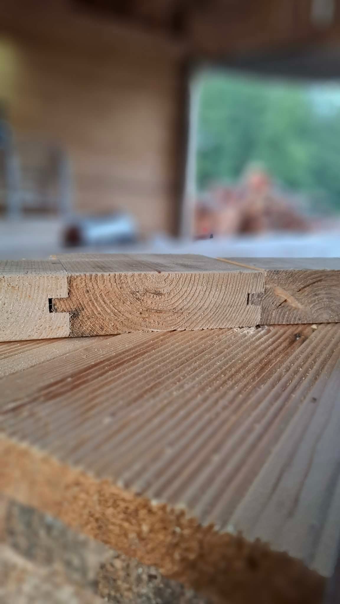 Połączenie na wpust i czop dwóch desek z widocznymi słojami drewna, w tle zamazane otoczenie warsztatu.