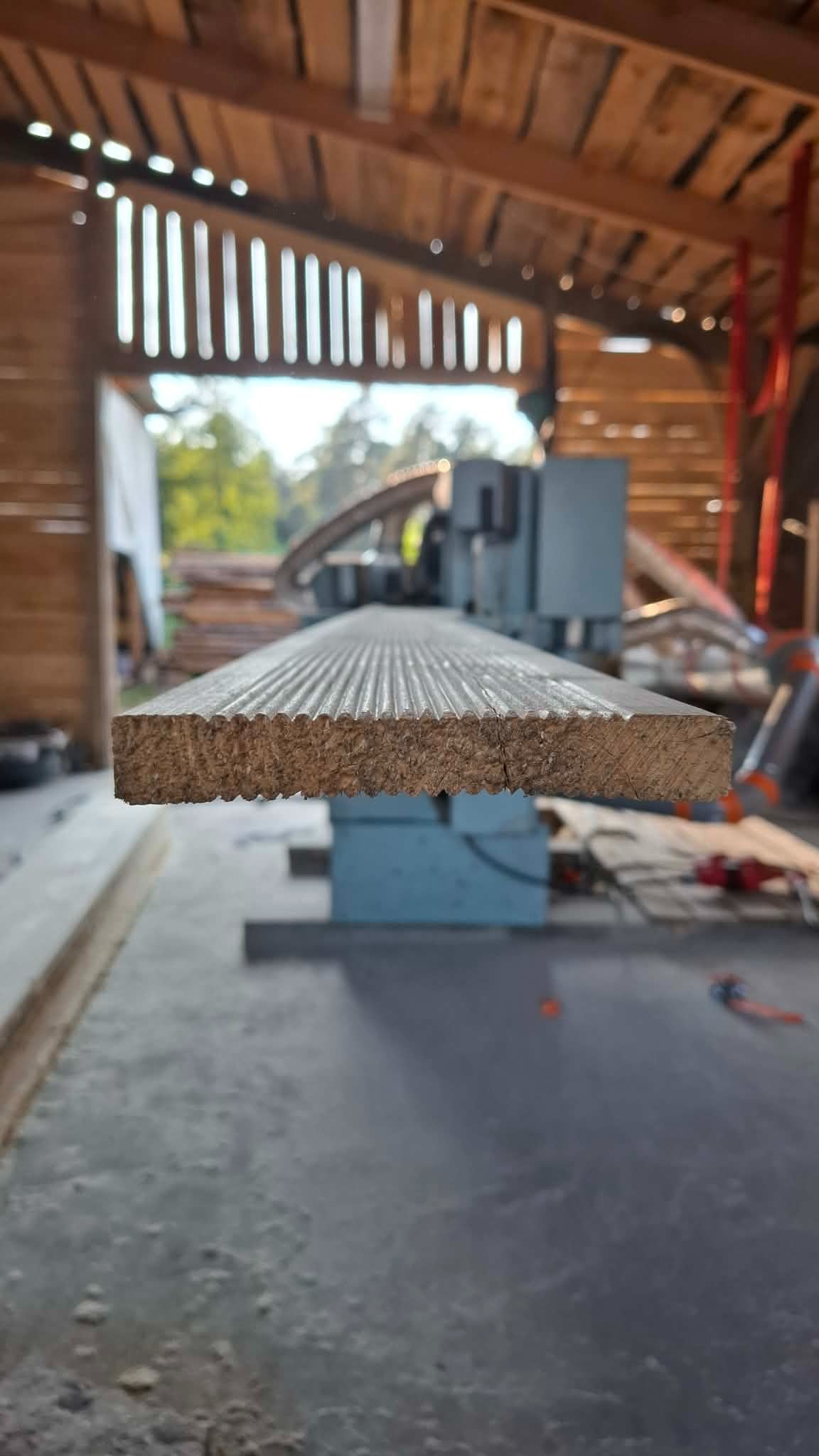 Profilowana deska z bliska, ukazująca teksturę i wzór, na tle hali z maszyną do obróbki drewna. Oświetlenie naturalne, surowy wygląd.