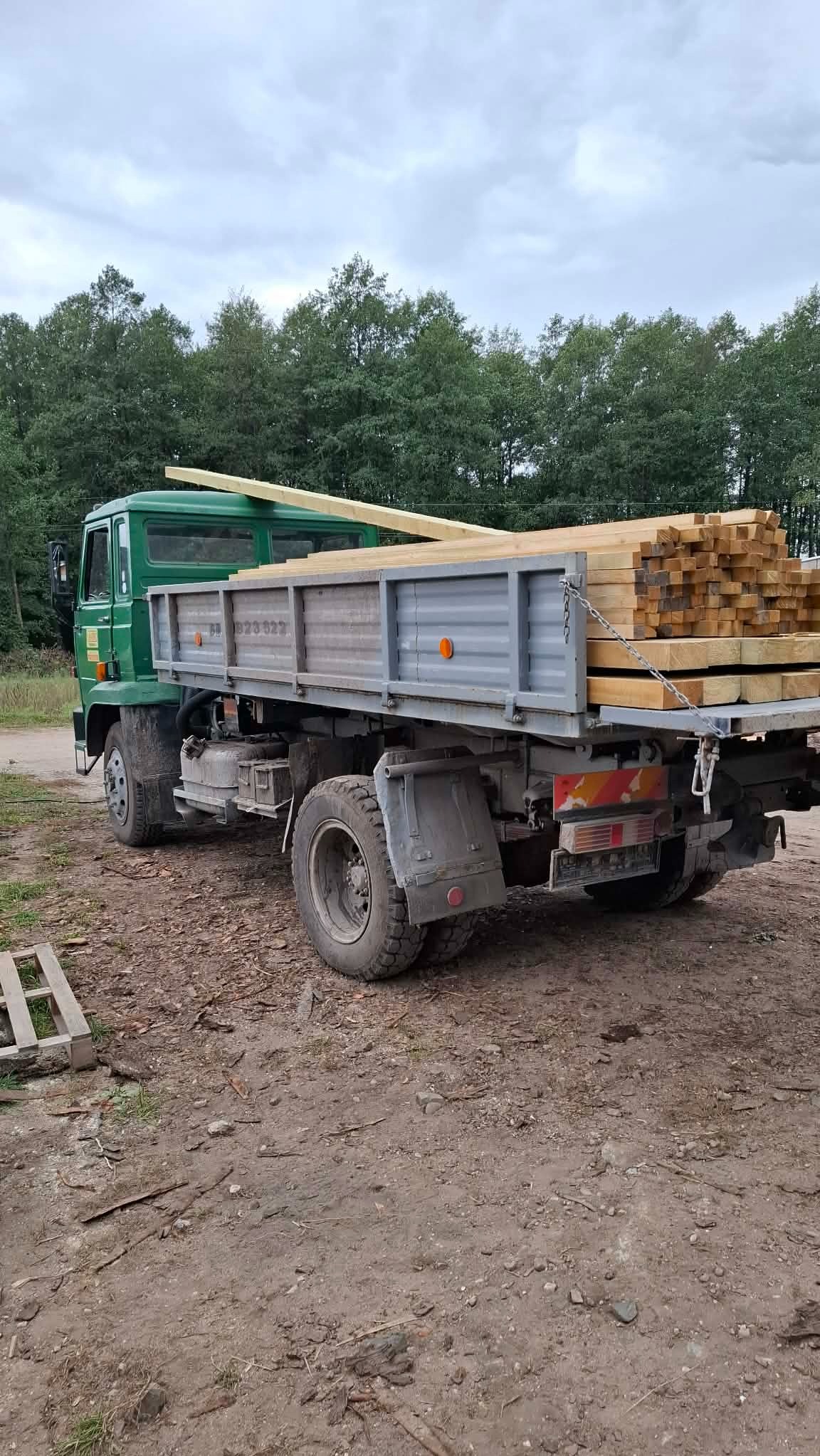 Ciężarówka z ładunkiem świeżo przetartego drewna na pace, zabezpieczonego łańcuchami, na tle zieleni drzew. Widoczne koła i fragment palety.