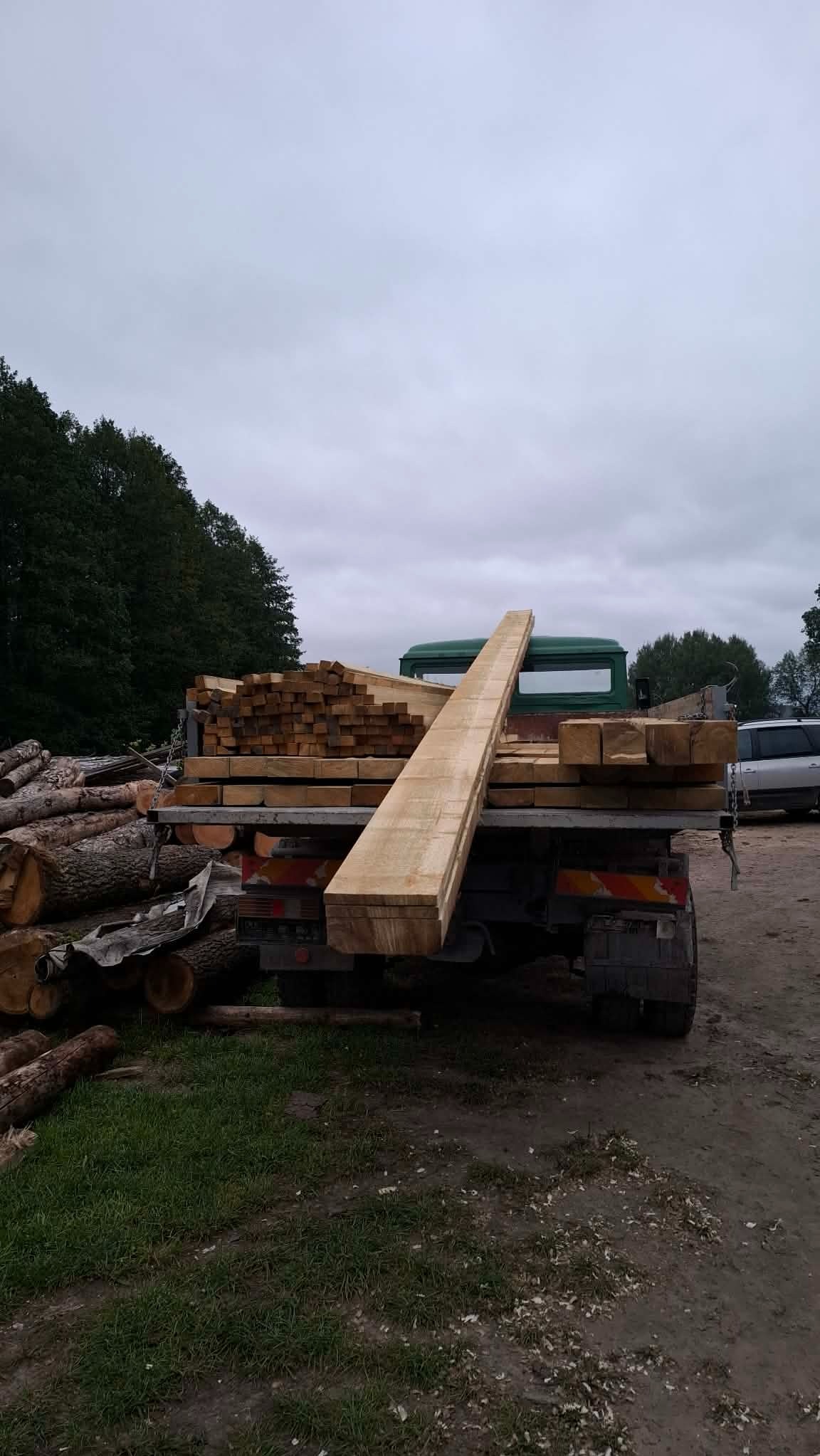 Ciężarówka z ładunkiem świeżo przetartego drewna, w tle stosy pni drzew i pochmurne niebo. Widoczne ślady obróbki drewna.