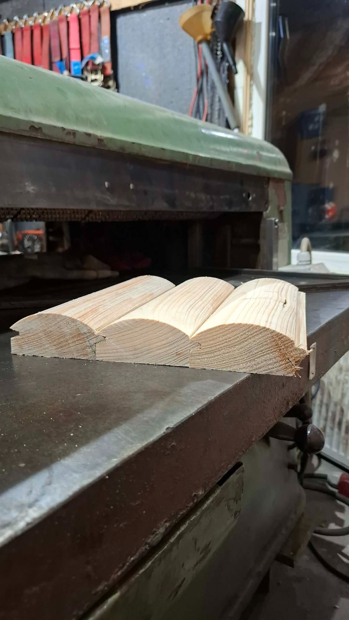 Profilowane listwy z jasnego drewna leżą na metalowym stole roboczym, prawdopodobnie po obróbce na stolarce. Widoczne ślady obróbki i naturalna struktura drewna.