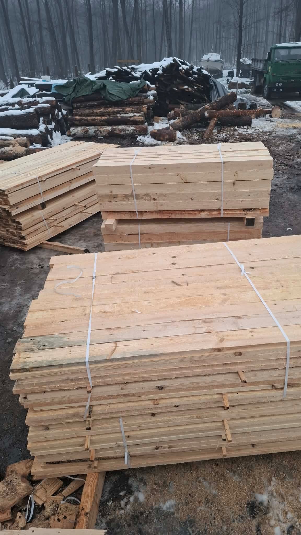 Stosy jasnego drewna budowlanego, spięte białymi taśmami, na tle zimowego krajobrazu z pniem drzewa i zielonym samochodem ciężarowym w oddali.