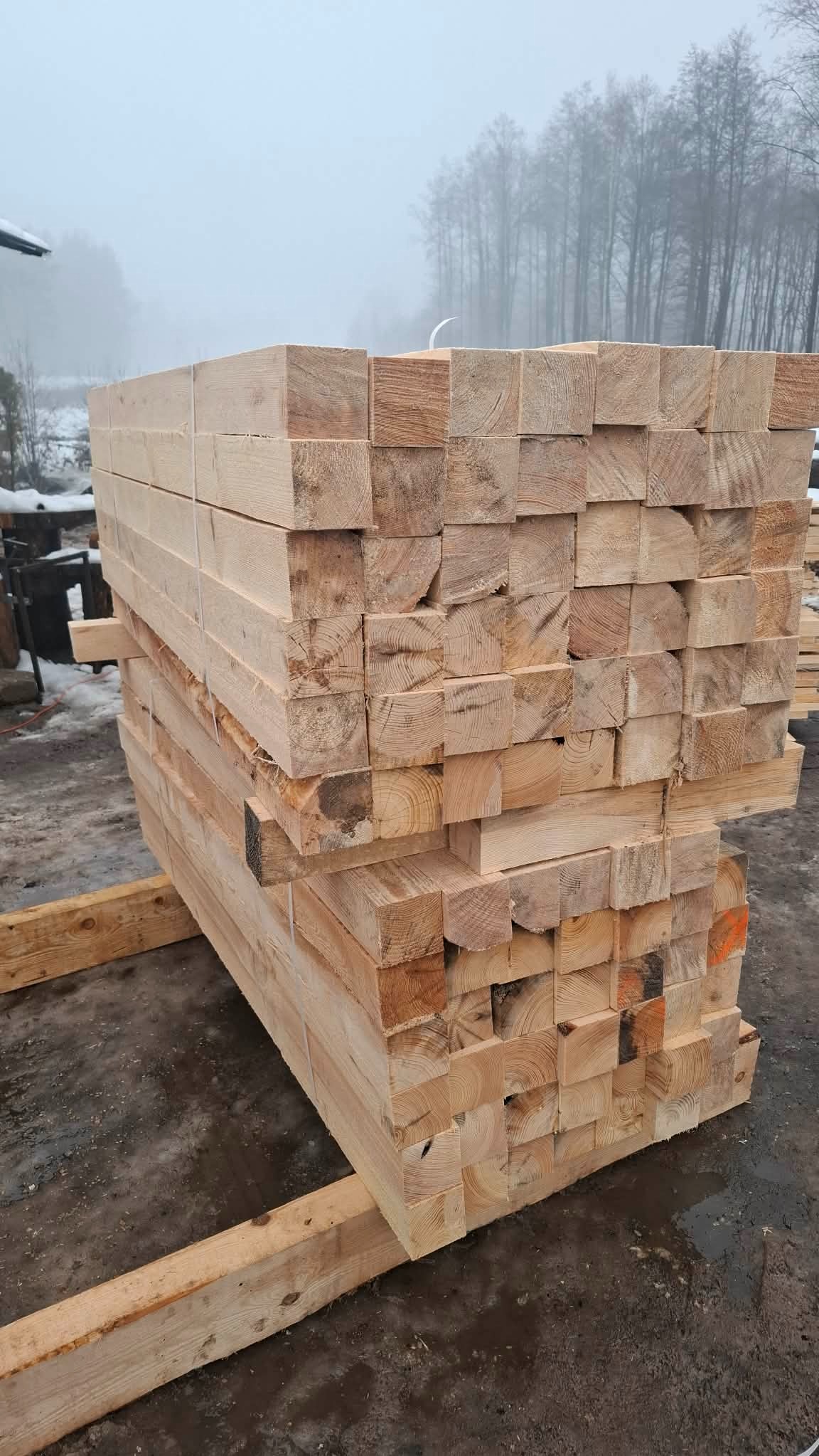 Stos belek z jasnego drewna ułożonych na legarach, widoczne słoje drewna, tło z drzewami we mgle, zimowa sceneria. Drewno budowlane przygotowane do transportu.