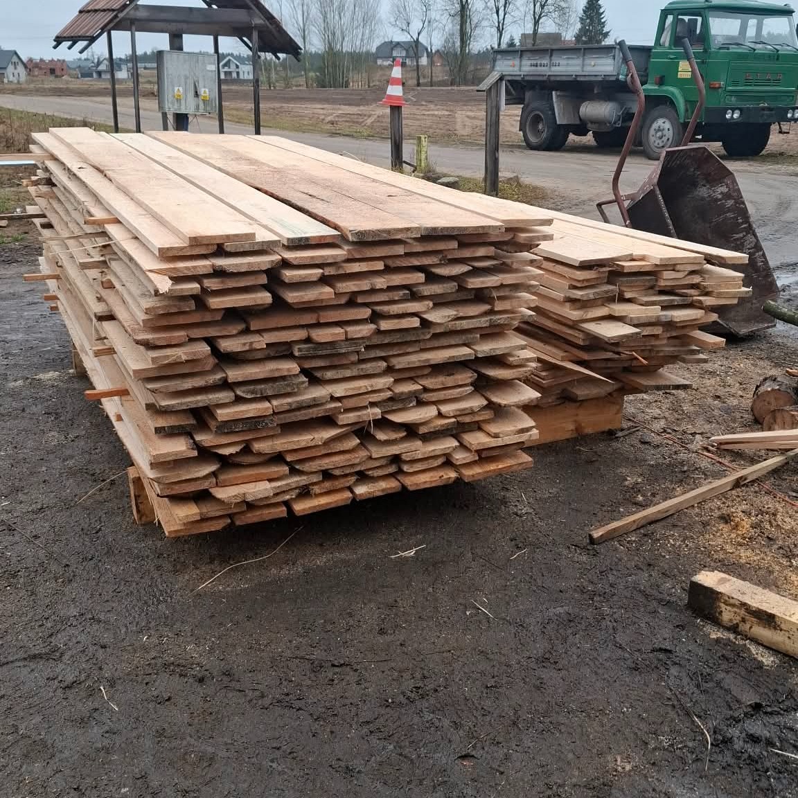 Stosy świeżo przetartego drewna na paletach, z widocznym zielonym samochodem ciężarowym w tle, sugerujące skład drewna lub przygotowanie do budowy.