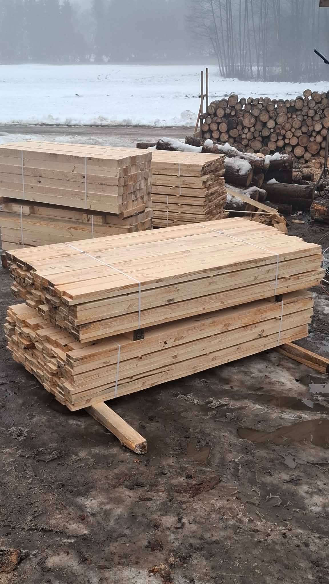 Stosy świeżo przetartego drewna budowlanego, zabezpieczone sznurkiem, składowane na zewnątrz w zimowych warunkach. W tle stosy pociętych pni drzew.