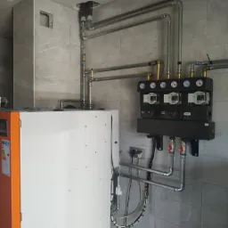 Pomarańczowo-biały piec z podłączonymi rurami i czarnym rozdzielaczem z manometrami na tle szarych płytek, ręka dotyka ekranu pieca.