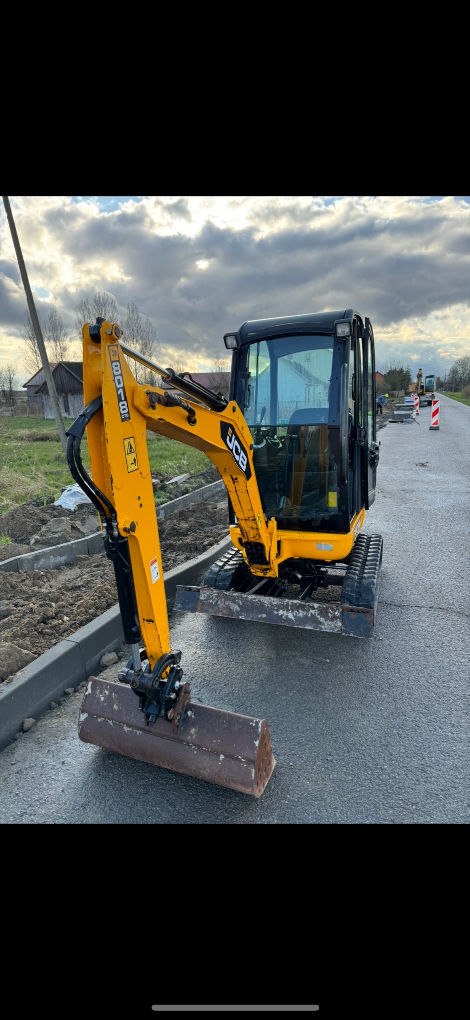 Żółta mini koparka JCB na drodze z nowymi krawężnikami i kostką w tle, pochmurne niebo.