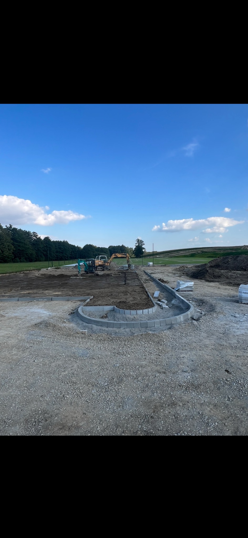 Przygotowanie terenu pod ścieżkę z obrzeżami betonowymi, widoczne maszyny budowlane na tle zieleni i błękitnego nieba.