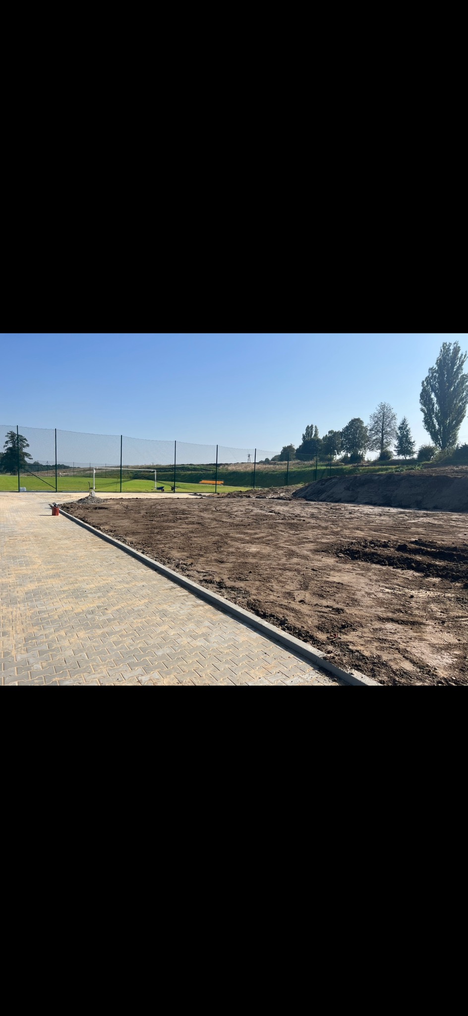 Przygotowany teren pod przyszłą inwestycję, częściowo wyłożony kostką brukową, z widoczną ziemią i siatką ogrodzeniową w tle.