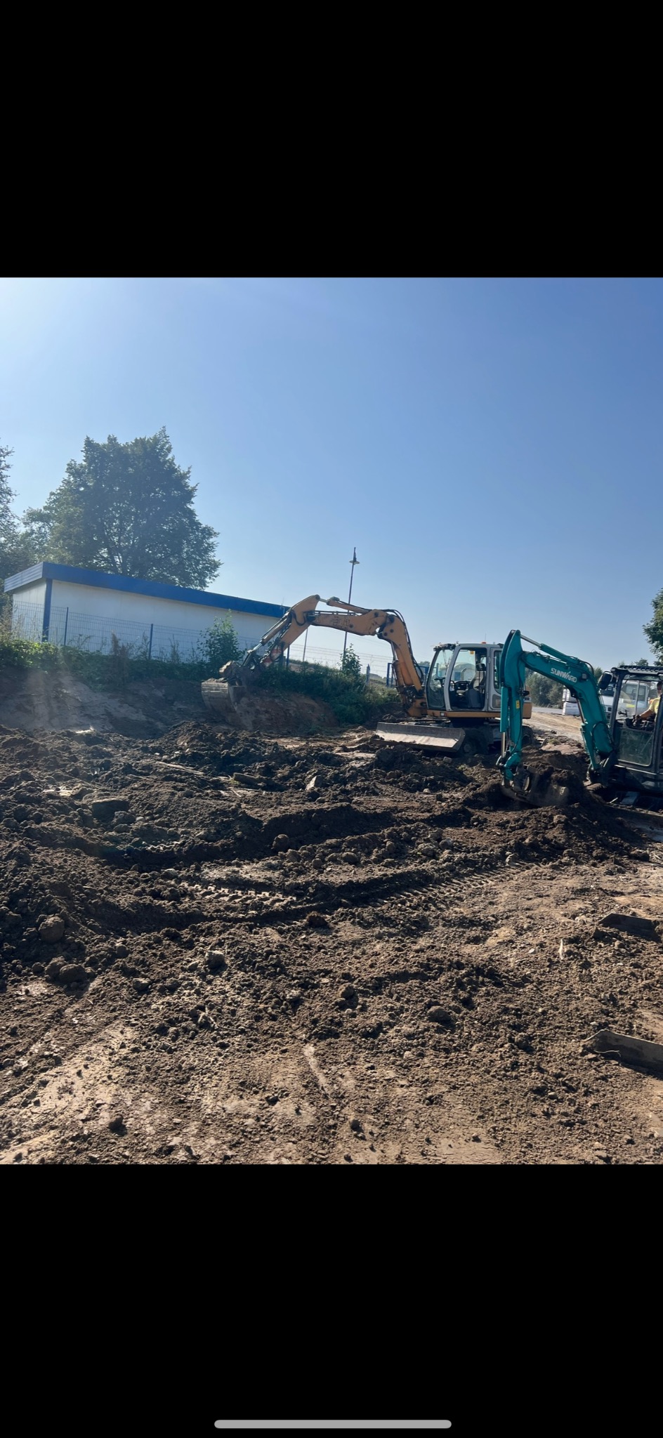 Żółta koparka w trakcie prac ziemnych na placu budowy, obok zielona mini koparka, w tle budynek z białymi ścianami i niebieskim dachem, teren nierówny, pokryty błotem.