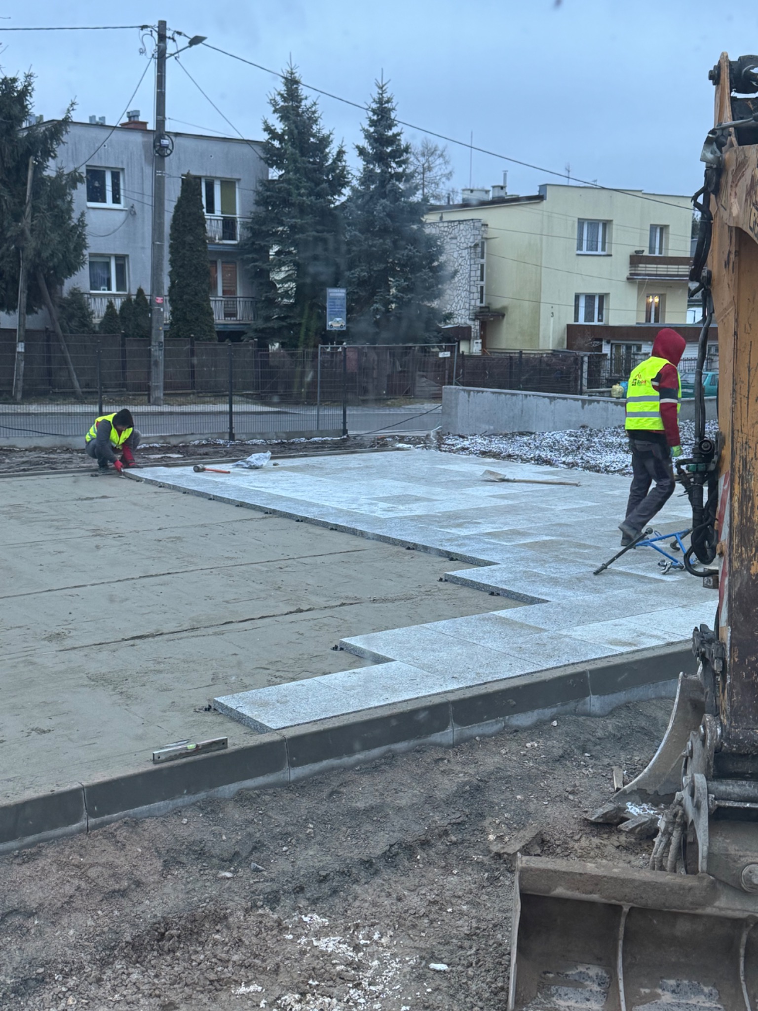 Układanie jasnoszarej kostki granitowej na przygotowanym podłożu, z widocznym fragmentem koparki i pracownikami w odblaskowych kamizelkach w tle zabudowy miejskiej.