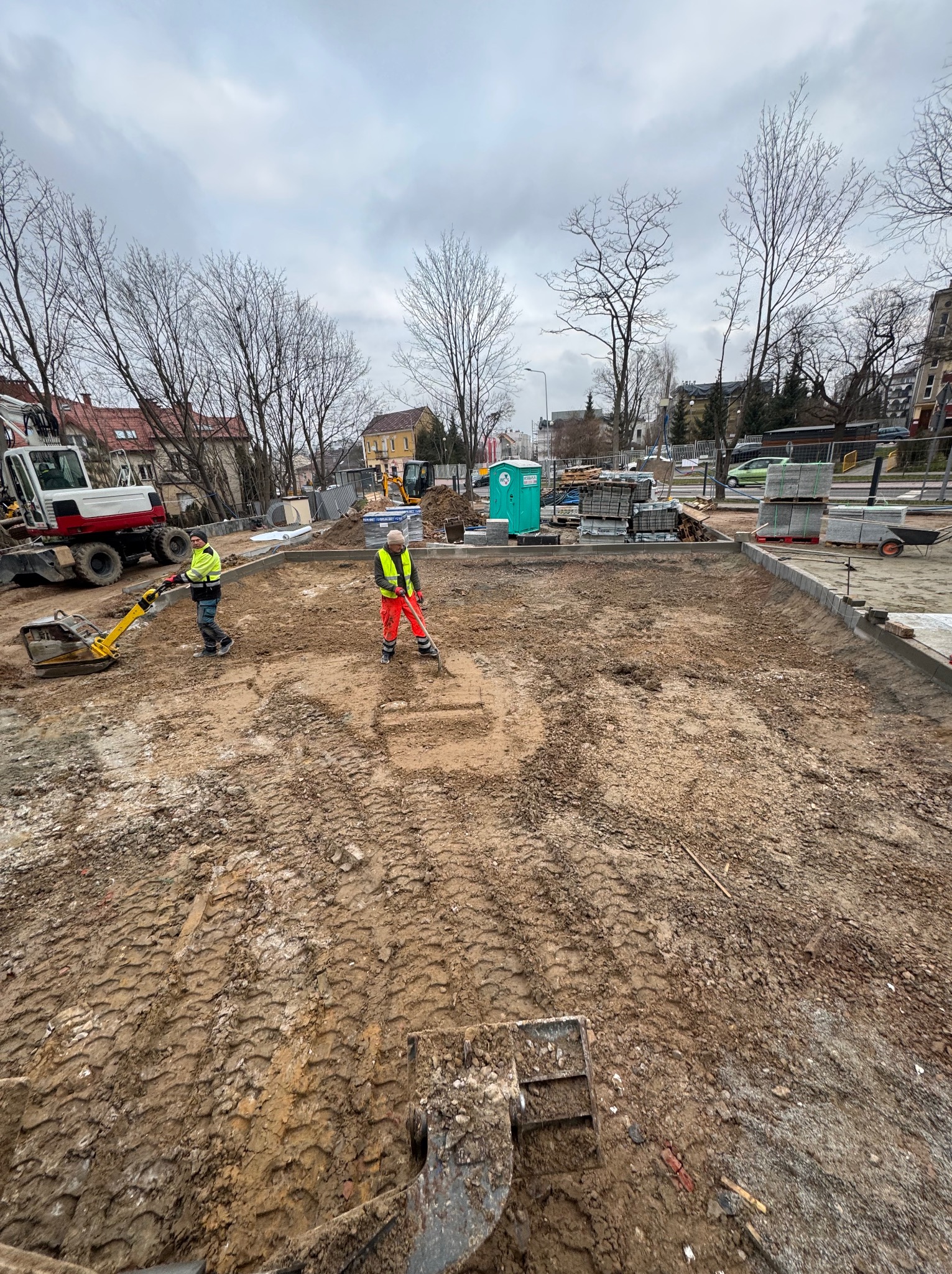Przygotowanie terenu pod budowę: dwóch pracowników w odblaskowych ubraniach, jeden ubija ziemię zagęszczarką wibracyjną, drugi wyrównuje teren łopatą, widoczne ślady opon i maszyny budowlane...