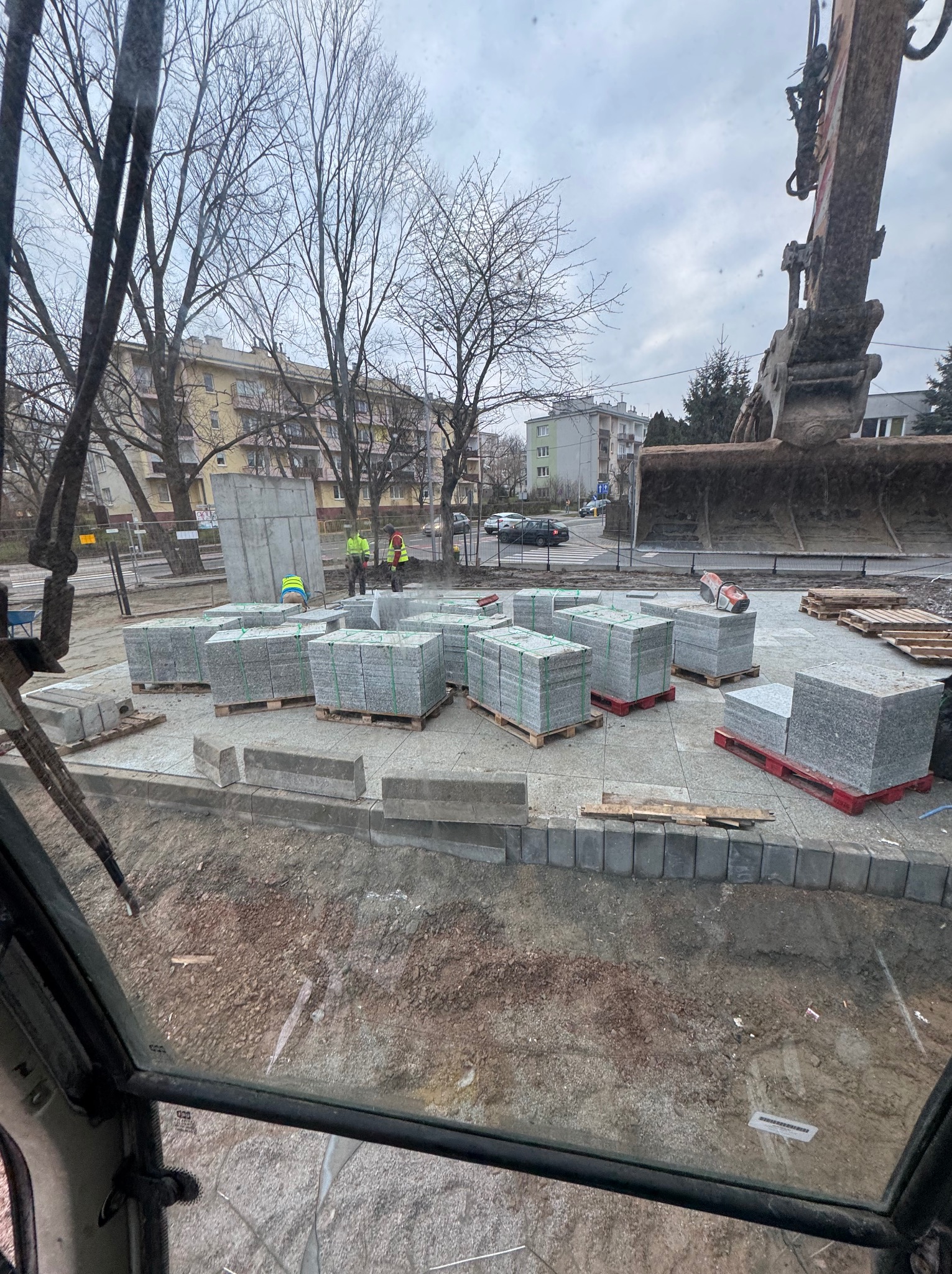 Widok z kabiny koparki na plac budowy: przygotowane palety z kostką granitową, robotnicy w kamizelkach odblaskowych, fragment wykopu i zabudowa miejska w tle.