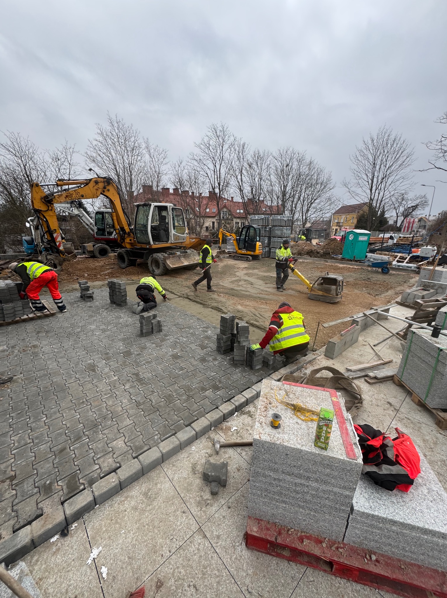 Ekipa budowlana w trakcie układania szarej kostki brukowej na dużej powierzchni, z widocznymi maszynami budowlanymi w tle i stertami kostki do ułożenia