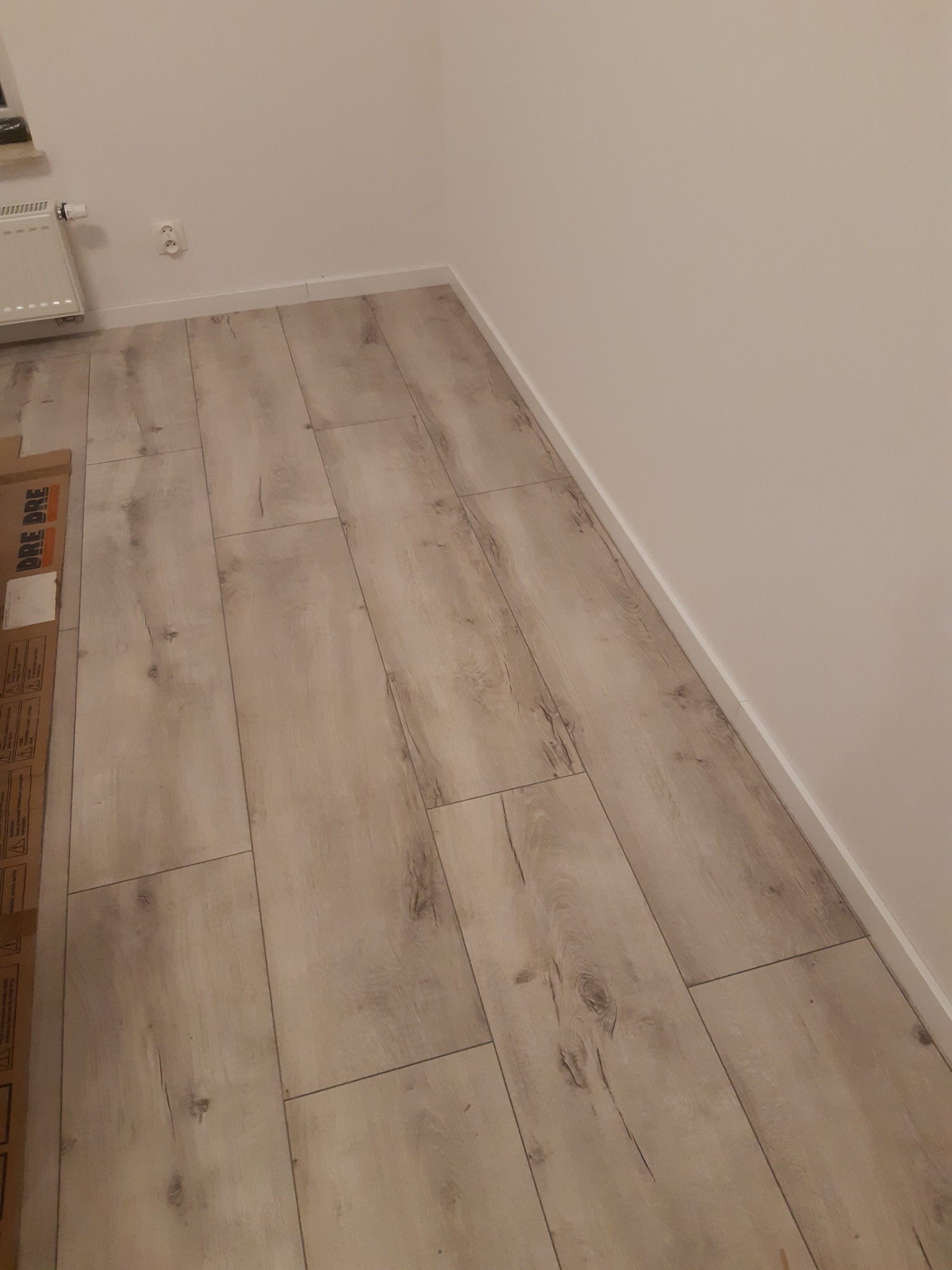 Ułożona podłoga z paneli imitujących jasne drewno, widoczna przy ścianie z białą listwą przypodłogową, fragment kartonu z oznaczeniami producenta w lewym dolnym rogu.