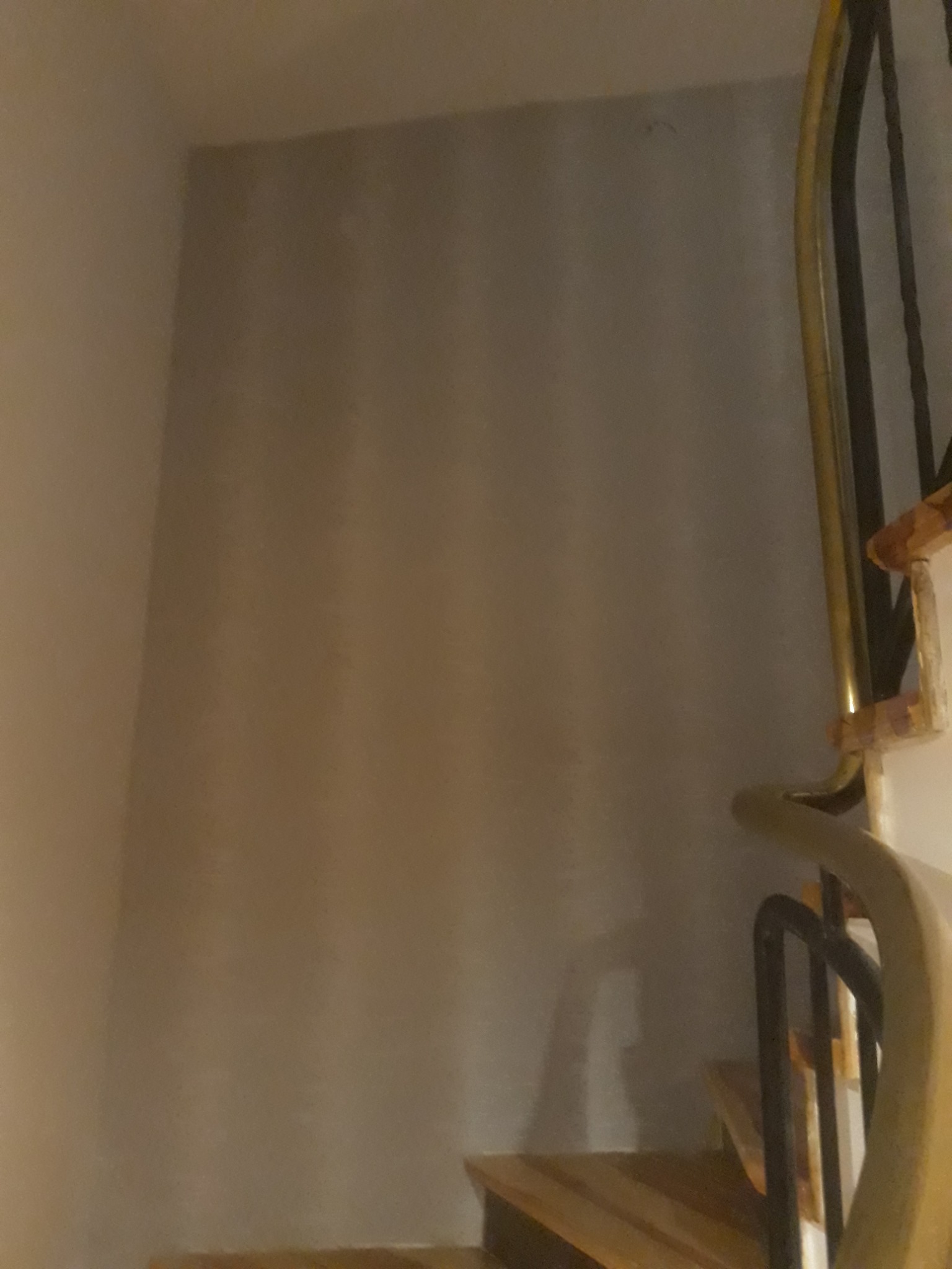 Ściana pokryta tapetą w odcieniach szarości, widoczna obok fragmentu schodów z drewnianymi stopniami i metalową balustradą z elementami w kolorze złotym.
