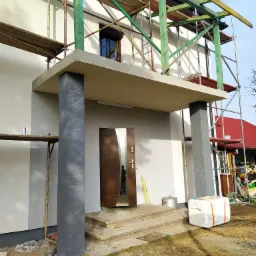 Dom jednorodzinny w trakcie prac elewacyjnych: ocieplanie z użyciem rusztowań, widoczne styropianowe płyty, świeżo pomalowane kolumny wejściowe w kolorze grafitowym, częściowo odsłonięte wejście.