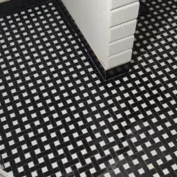 Podłoga w łazience wyłożona czarno-białą mozaiką w wzór koszykowy, obok fragmentu ściany wyłożonej białymi, prostokątnymi płytkami, widoczne ślady prac remontowych.