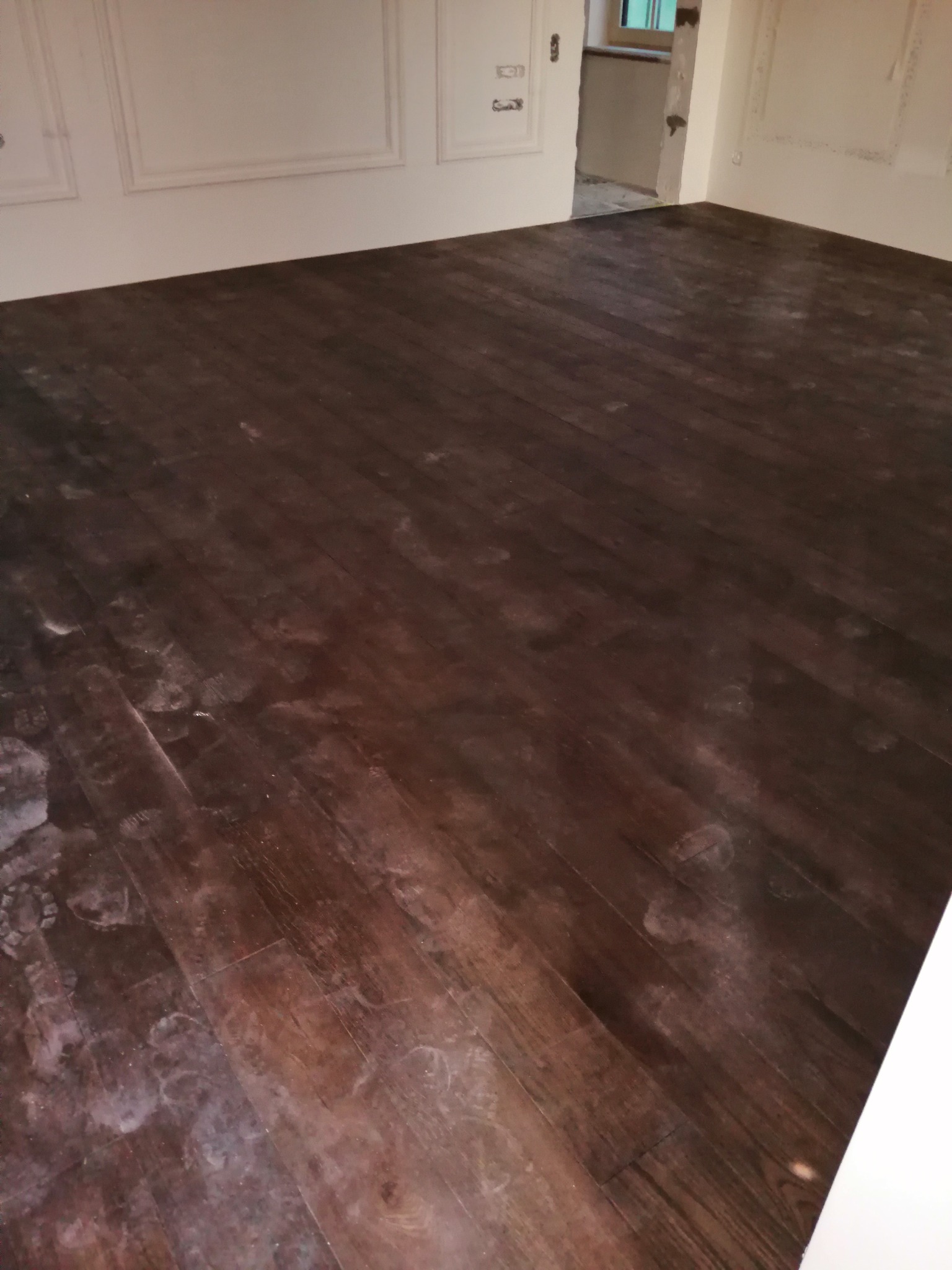 Ciemny, drewniany parkiet z widocznymi śladami białego pyłu lub kurzu, prawdopodobnie po szlifowaniu lub innym procesie obróbki, w pomieszczeniu z białymi ścianami i dekoracyjnymi panelami.