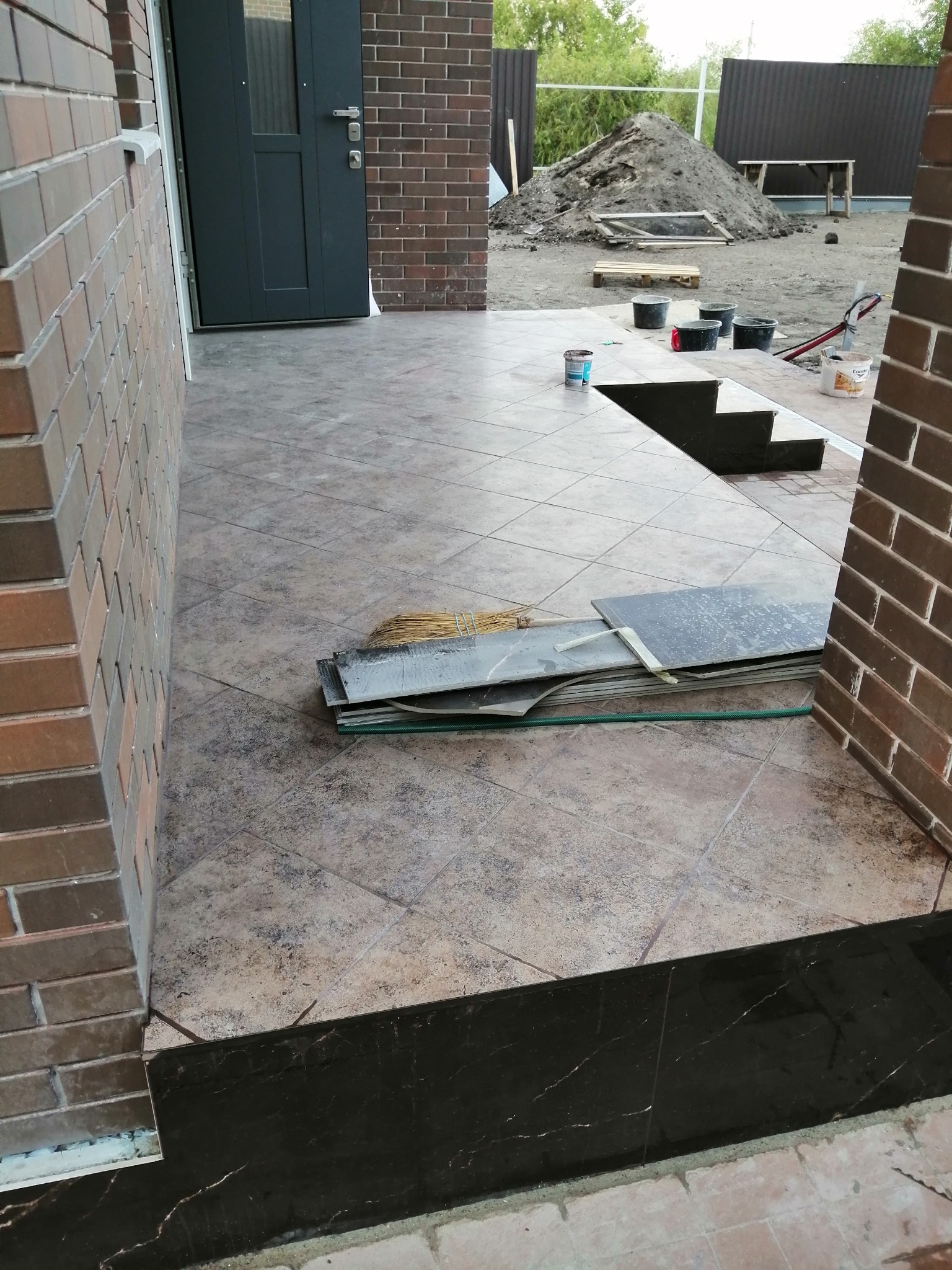 Ułożona beżowa terakota na zewnątrz domu z widocznymi narzędziami i pozostałościami po pracy, w tle sterta ziemi.