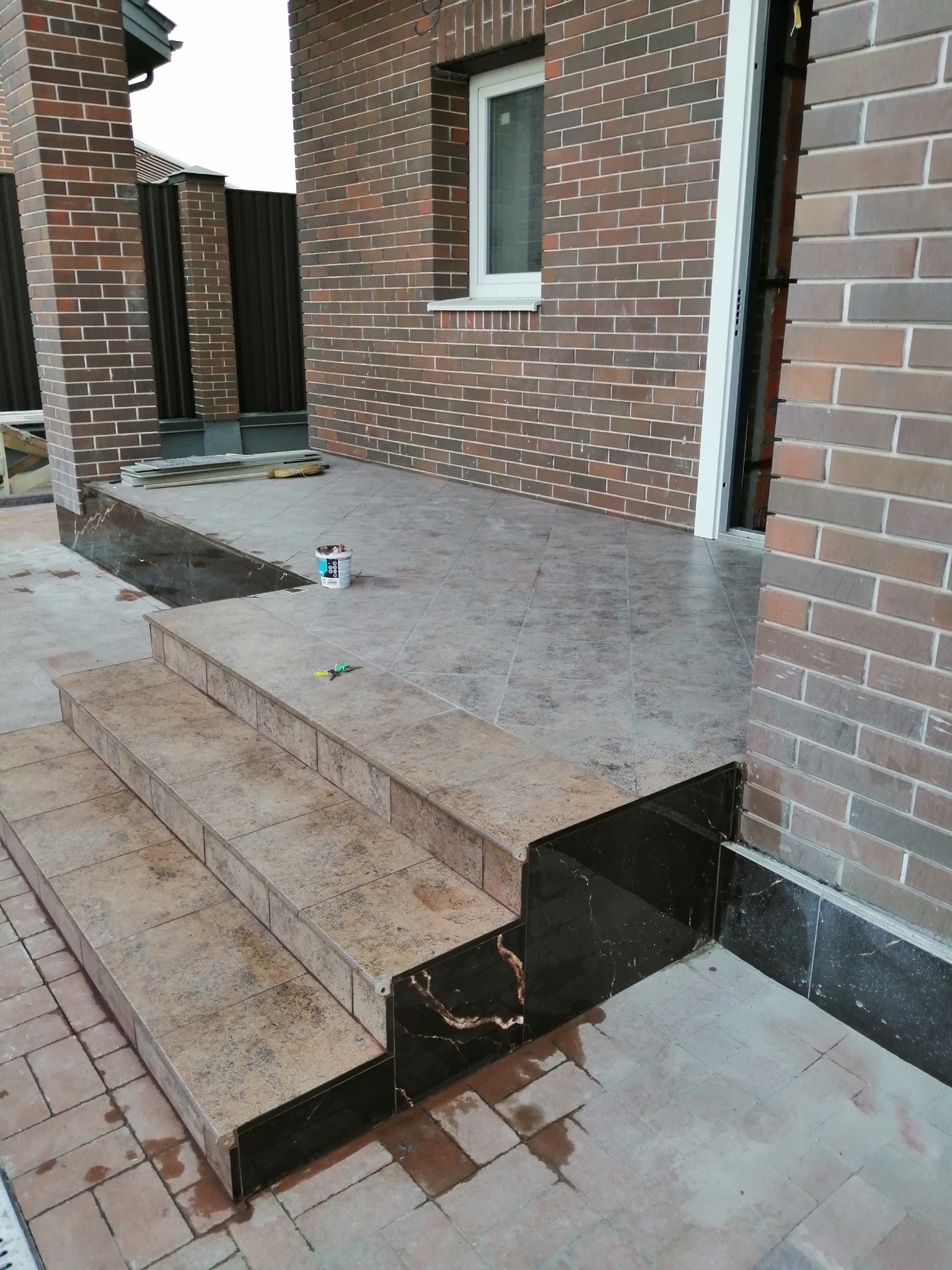Wykończone schody wejściowe do domu z brązowej cegły, płytki ceramiczne na stopniach i czarne marmurowe obłożenie boczne, widoczny fragment tarasu z płytkami, puszka farby i narzędzia na górnym...