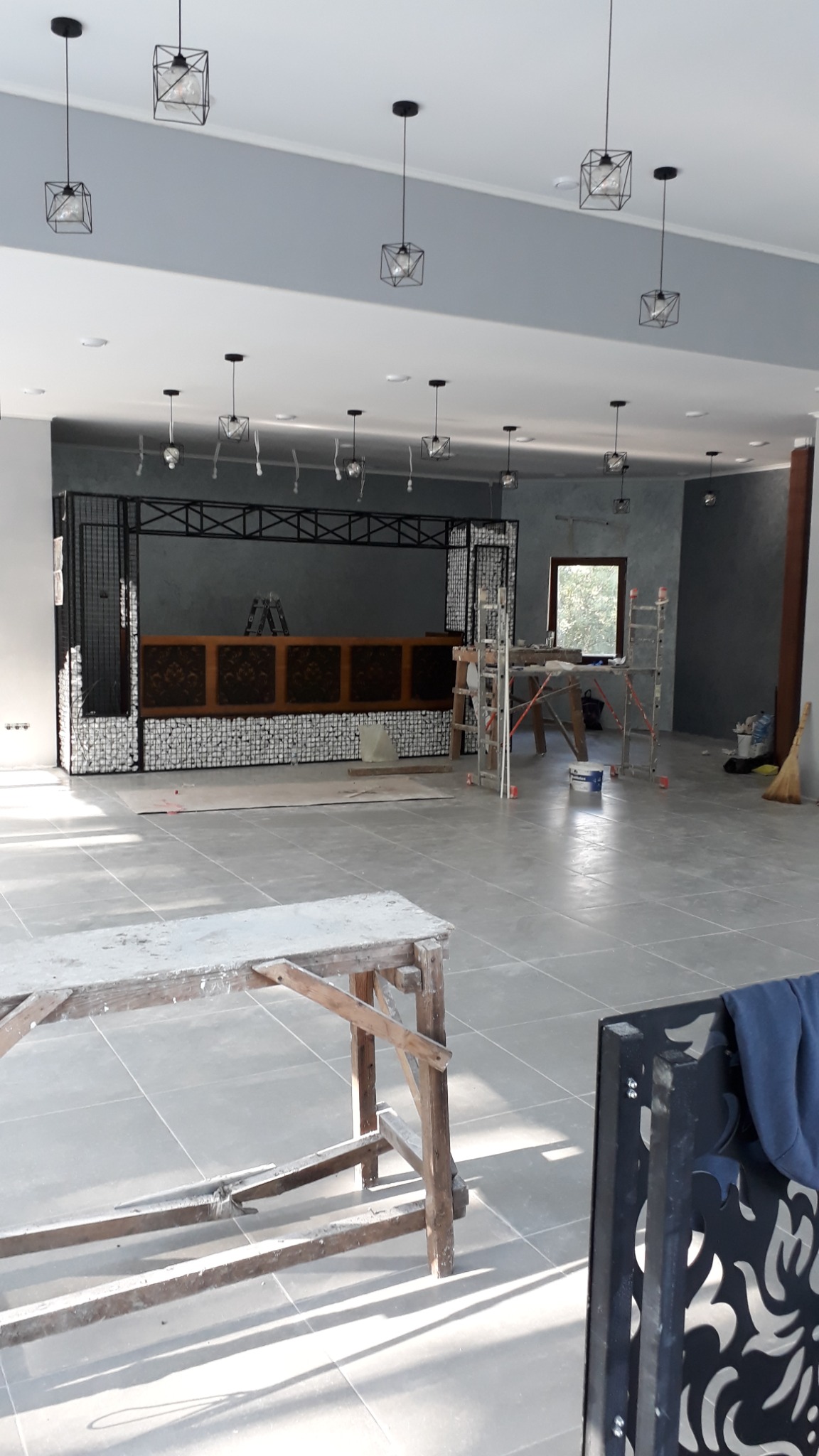 Duża sala w trakcie wykończenia z szarymi ścianami, metalową konstrukcją baru z drewnianymi elementami, licznymi lampami w kształcie klatek i drewnianym stołem malarskim na pierwszym planie.